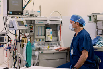 A color photograph of a modern surgical room with a medical professional in blue scrubs and a surgical mask seated next to an anesthesia machine and various monitors. The equipment includes a Philips monitor and a Datex-Ohmeda anesthesia machine, highlighting the advanced medical technology used during surgeries.