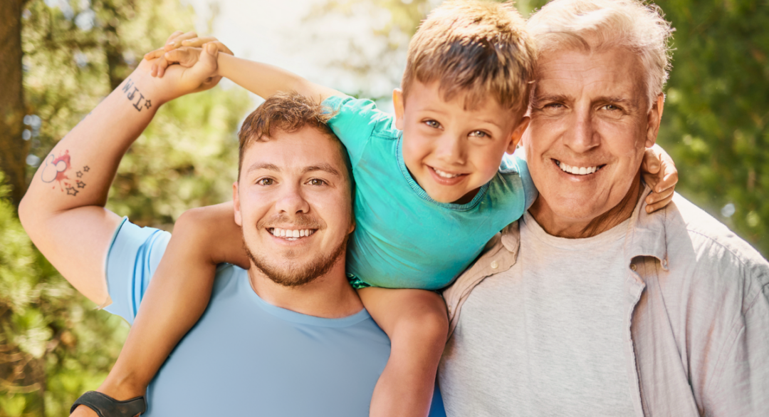 Three generations of men outdoors smiling, featuring a young boy on the shoulders of a man with tattoos, and an elderly man beside them.