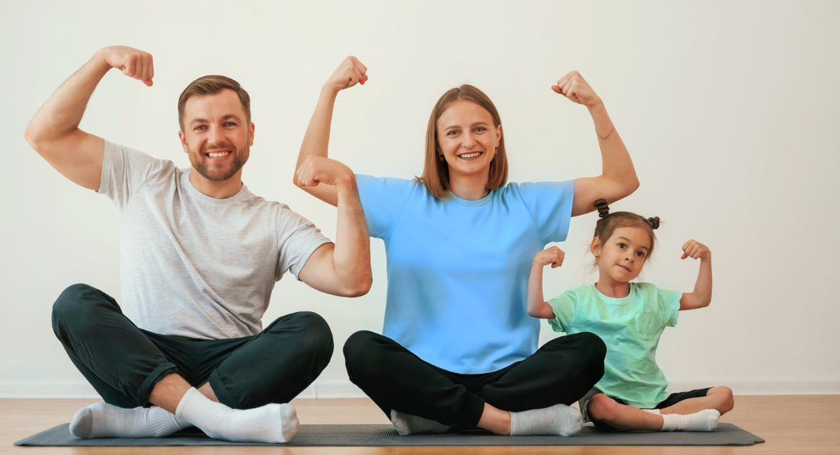 A family sitting on the floor, flexing their muscles. The father, mother, and young daughter are all smiling and showing their biceps in a playful and fun atmosphere.