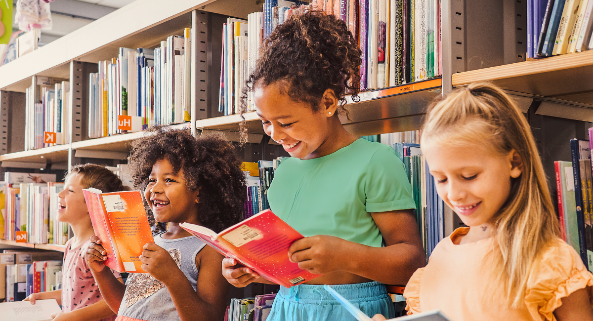 A group of four children, diverse in ethnicity, are standing in a library, joyfully reading books together and smiling