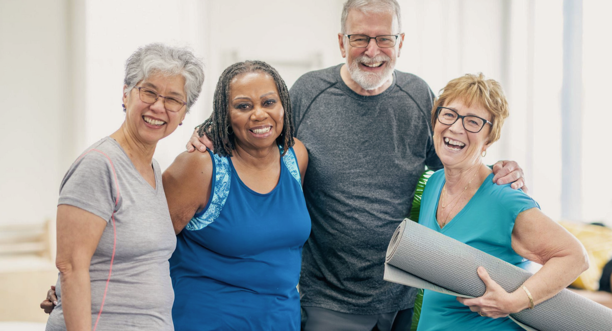 A group of four senior adults, two women and two men, smiling and standing close together in a bright, indoor setting. One woman is holding a yoga mat, indicating they are possibly participating in a wellness or fitness activity.