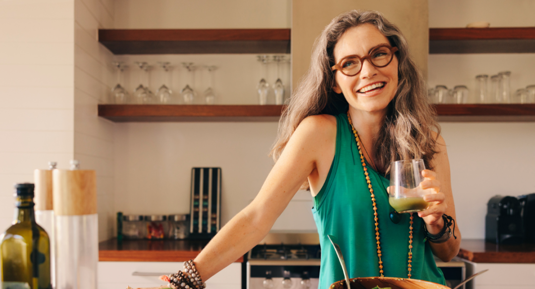 A woman with long gray hair and glasses smiles while standing in a modern kitchen. She is wearing a green sleeveless top and a beaded necklace, holding a glass of green juice. Shelves with glassware are visible in the background.