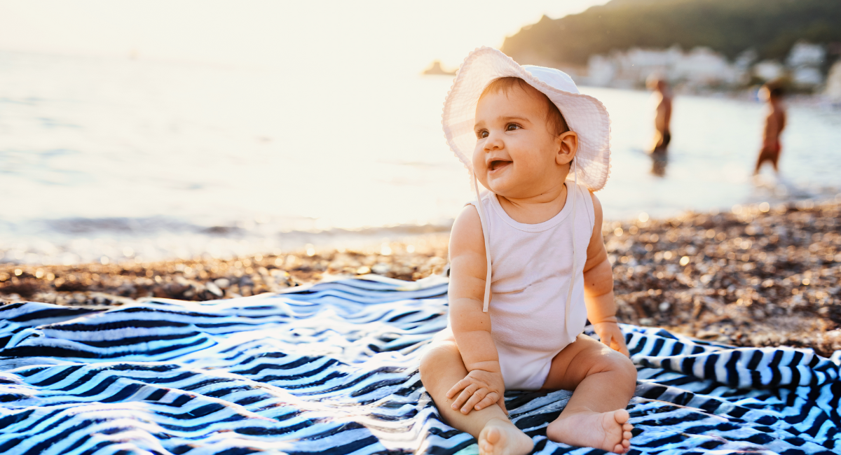 A baby in a white onesie and sun hat is sitting on a striped blanket at the beach, with a background of the sea and people in the distance