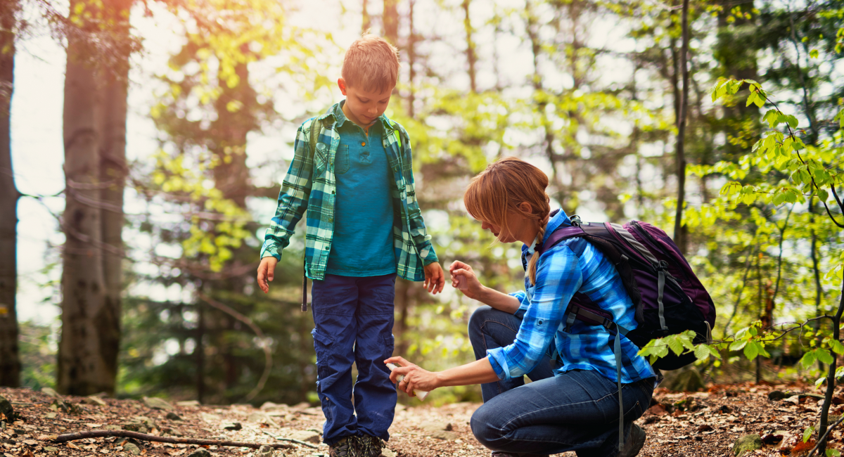 A parent kneels down to spray their child's pants while hiking in a sunlit forest. They are both wearing plaid shirts and backpacks