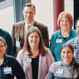 Group photo of Monadnock Community Hospital staff in the Wellness Center basement. From left to right: Armando Rangel, HR Generalist, in a plaid shirt and glasses; Cathy Maki, Accountant, in a teal sweater; Dr. Dmitry Tarasevich, Monadnock Internal Medicine, in a plaid jacket and tie; Tausey Wolfe, Monadnock Behavioral Health Practice Manager, in a cardigan; Corryn Nelson, Physical Therapist, in a black top; Jennifer Norris, Patient Access Coordinator, in a grey sweater; and Leah Hakala, Birthing Suite LNA, in blue scrubs.