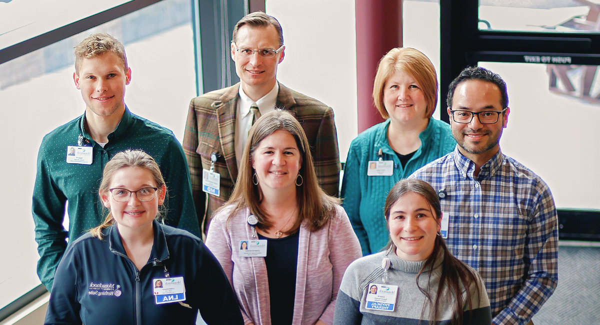 Group photo of Monadnock Community Hospital staff in the Wellness Center basement. From left to right: Armando Rangel, HR Generalist, in a plaid shirt and glasses; Cathy Maki, Accountant, in a teal sweater; Dr. Dmitry Tarasevich, Monadnock Internal Medicine, in a plaid jacket and tie; Tausey Wolfe, Monadnock Behavioral Health Practice Manager, in a cardigan; Corryn Nelson, Physical Therapist, in a black top; Jennifer Norris, Patient Access Coordinator, in a grey sweater; and Leah Hakala, Birthing Suite LNA, in blue scrubs.