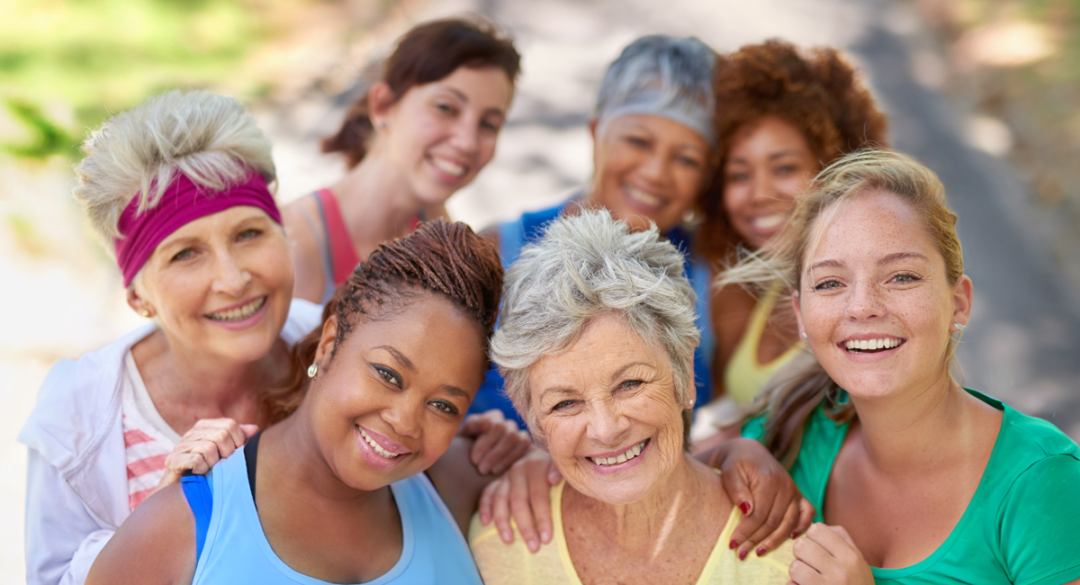 A diverse group of women of various ages and ethnicities smiling and posing together outdoors, showing a sense of community and friendship