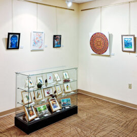 Art gallery wall displaying various paintings and artworks. A glass cabinet showcases framed illustrations, and colorful mandala artwork hangs on the walls