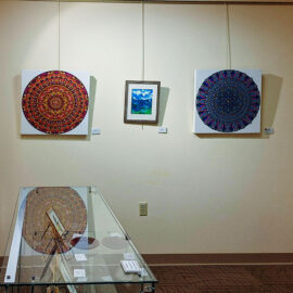 Gallery corner with vibrant mandala paintings and a mountain landscape. A glass cabinet displays framed art, creating an inviting and artistic atmosphere