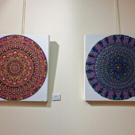 Two large mandala paintings displayed side by side in a gallery setting, showcasing intricate patterns and vibrant colors