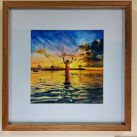 Framed watercolor painting titled 'Fisherman's Last Catch' by Amara Kanaparthi, depicting a fisherman casting a net in a tranquil water scene with a dramatic sunset and vibrant clouds in the background