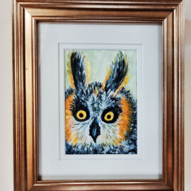 A close-up painting of an owl with wide, piercing yellow eyes. The owl's feathers are painted in shades of blue, orange, and gray, set against a soft, pale background. The painting is framed in a glossy gold frame