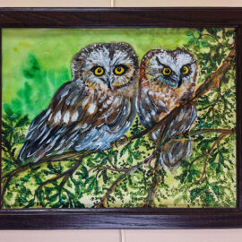 A close-up painting of two owls with bright yellow eyes, perched closely together on a branch. The owls are depicted with detailed brown and white feathers, set against a vivid green background. The painting is framed in a dark wooden frame and hung on a beige wall.