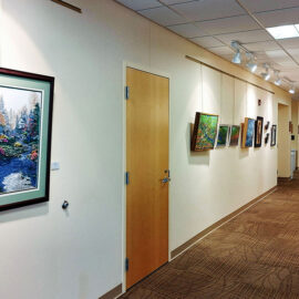 A long hallway of an art gallery, showcasing multiple paintings hung on both sides. The walls are beige, and the ceiling features overhead track lighting illuminating the artwork. The floor is carpeted in a neutral tone, creating a warm and inviting atmosphere. A quilt is displayed at the far end of the hallway