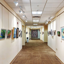 Hallway of an art gallery with paintings displayed on both sides of the wall. The right wall features various artworks, including a fish painting in the center, with spotlights highlighting each piece. The left side has additional paintings, showcasing diverse styles and subjects