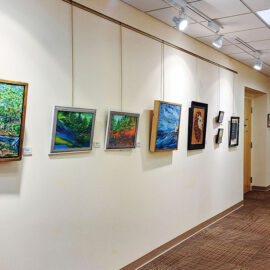 Another view of the art gallery hallway, displaying various paintings hung along the beige walls. The ceiling lights focus on the artwork, highlighting a diverse collection of landscapes and abstract pieces. The hallway is spacious and well-lit, with a quilt visible in the distance at the end of the corridor