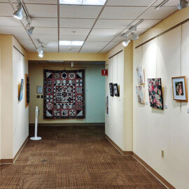 Art gallery corridor featuring various paintings on display. The right wall showcases several colorful artworks, including floral paintings and portraits. The left wall displays more abstract pieces, with a large quilt hanging at the end of the hallway, adding texture and depth to the scene