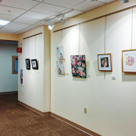 Art gallery hallway showcasing a variety of paintings on both walls. The right wall features floral and portrait artworks, while the left wall displays abstract and landscape pieces. A quilt with intricate patterns hangs at the far end of the hallway, adding a traditional touch to the modern display