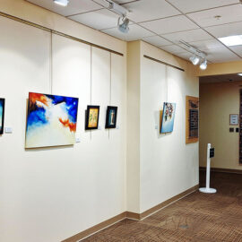 Hallway in an art gallery, highlighting paintings of abstract and colorful designs on the left wall. The right wall features several floral and portrait artworks, with spotlights illuminating each piece. A large quilt with detailed patterns is displayed at the end of the hallway