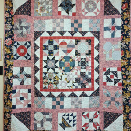 A quilt featuring intricate patchwork patterns with floral borders and a central geometric design, incorporating colors like pink, navy, and white