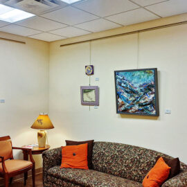 A corner of a family room showcasing multiple artworks, including landscapes and abstract pieces, hung above a patterned couch with colorful cushions