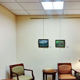 Cozy corner with a small gallery wall in a waiting room at Monadnock Community Hospital. Features a single painting of a mountain landscape on the left wall and two smaller paintings on the center wall above a pair of armchairs and a small table with a book. A lamp is on the right, providing warm light, enhancing the comfortable atmosphere