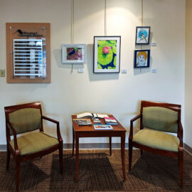 Two armchairs positioned around a small table in the waiting area at Monadnock Community Hospital. Above, a display of colorful paintings and drawings, including a vibrant flower and birds, adorns the wall. The hospital's directory sign is prominently featured next to the artwork