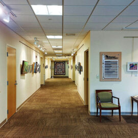 A hallway corner at Monadnock Community Hospital featuring a series of framed artworks hanging along the wall. The art ranges from colorful landscapes to abstract designs, creating a welcoming and engaging environment. Two chairs and a table with brochures are placed in front of the art wall