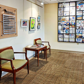 The corner of a waiting area at Monadnock Community Hospital, showcasing a large wall mural composed of many small, colorful tiles. Two chairs with a table filled with informational pamphlets are positioned to the left of the mural. Above the chairs, a series of framed artworks hang on the wall