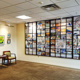 A wider view of the waiting area at Monadnock Community Hospital, highlighting a large, vibrant tile mural covering an entire wall. On the adjacent wall, a series of framed artworks hang above two chairs and a table, creating a colorful and artistic atmosphere in the hallway