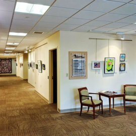 A hallway view at Monadnock Community Hospital, showing artwork displayed on both sides. The left side features a long row of framed paintings and drawings, while the right side displays a tile mural and additional art pieces. The floor is carpeted, contributing to the warm and welcoming environment.