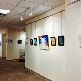 A hallway at Monadnock Community Hospital showcasing a collection of framed artworks on the wall. The artwork includes both colorful and monochromatic pieces, each with a small information card. The hallway is brightly lit, emphasizing the art and creating a gallery feel