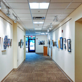 A long hallway in Monadnock Community Hospital featuring an array of framed artworks. The paintings, hung at eye level, offer a mix of vibrant colors and serene landscapes, enhancing the welcoming and calming atmosphere of the hospital's interior spaces