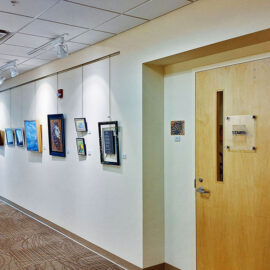 A hospital corridor at Monadnock Community Hospital, featuring an array of framed artworks displayed along the wall. The art includes vibrant abstract and nature-themed pieces. A door labeled 'Stairs' is visible on the right, with a small framed piece featuring an owl next to it