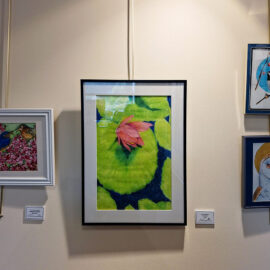 A close-up view of three framed artworks at Monadnock Community Hospital. The left piece depicts two colorful birds among blossoms. The central piece is a large green and pink abstract artwork resembling a lotus leaf and flower. The right piece shows a bird perched on a branch, and below it, a portrait of an owl with a golden halo