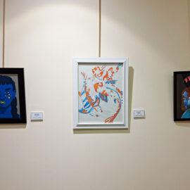 Three framed artworks on display at Monadnock Community Hospital. The left and right artworks are portraits with a blue theme, featuring stylized faces. The central piece is an abstract depiction of a fish in motion, using vibrant orange and blue colors