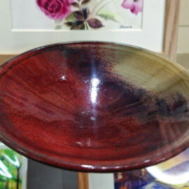 A ceramic bowl by Jenny Scholl, showcasing Japanese-influenced design with a deep red and purple glaze on the inside. The bowl features a glossy finish, creating a striking and elegant piece, displayed in a glass cabinet against a backdrop of floral artwork