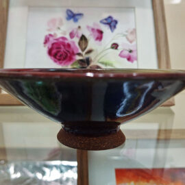 A side view of a ceramic bowl by Jenny Scholl, highlighting its smooth, curved design with a dark glossy exterior. The bowl sits on a small foot, and the background features delicate floral artwork framed in wood