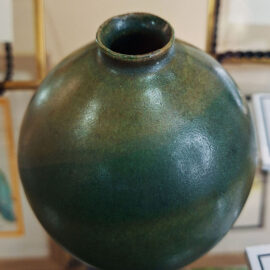 A ceramic orb by Jenny Scholl, influenced by Japanese design, featuring a smooth, matte green glaze. The orb is displayed on a stand, emphasizing its round shape and artistic simplicity against a background of framed artwork