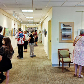 A group of people attending an art exhibition at Monadnock Community Hospital. Visitors are seen appreciating the artwork displayed along the corridor walls, with vibrant paintings and intricate designs creating an engaging environment