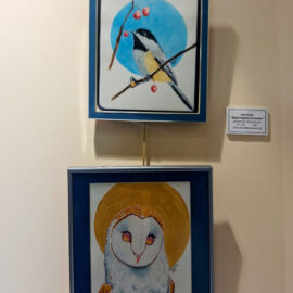 A pair of watercolor paintings by Kiera Burak, displayed vertically. The top piece shows a black-capped chickadee on a branch with a blue circular background, while the bottom piece features an owl with a golden halo against a deep blue frame