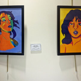 Two framed paintings side by side. The left painting features a woman with blue and green hues across her face, gazing upward against a purple background. The right painting depicts a woman with deep blue hair and vibrant orange skin, her lips are painted a striking blue, and the background is a bold yellow