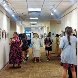 A hallway art gallery with visitors viewing various paintings displayed on the walls. People are casually dressed, examining the artwork as they move through the corridor, which is well-lit with overhead lights