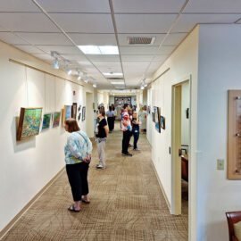 A photograph of an art gallery hallway with framed artworks displayed on both sides of the walls. Several people are admiring the art, with a clear view of the gallery's well-lit interior. The foreground shows a woman wearing a floral dress holding a phone, while others are scattered throughout the hallway, viewing the paintings