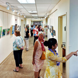 A wider view of the art gallery hallway, showing more people examining the artwork. The corridor is filled with visitors, some standing close to the paintings while others converse. A wall directory with the Monadnock Community Hospital logo is visible on the right, indicating the location of various hospital facilities