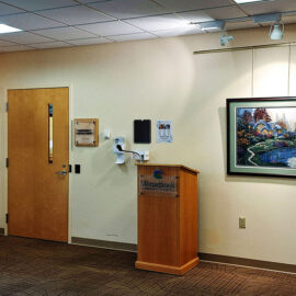 A corner of the gallery at Monadnock Community Hospital, showcasing a few framed artworks. On the left, a painting of cows is displayed, while a serene landscape painting hangs on the right. A podium stands between the artworks, creating a welcoming space for speeches or presentations. The area is well-lit, emphasizing the vibrant colors of the paintings