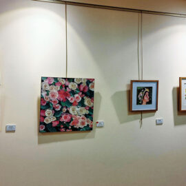 A section of a gallery wall displaying six paintings, including two square floral canvases and several framed portraits and floral artworks. The pieces are displayed on a beige wall, illuminated by gallery lighting