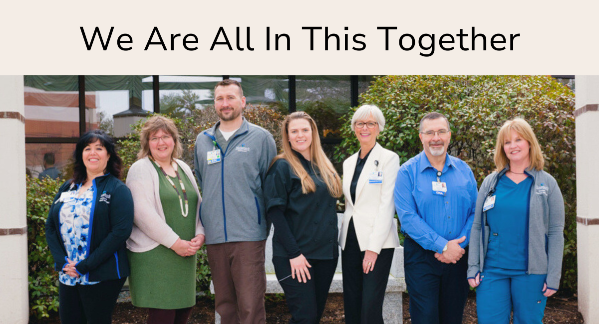 A group of seven Monadnock Community Hospital employees standing in front of the hospital building. They are positioned outdoors, dressed in a mix of professional and casual attire. The text above them reads 'We Are All In This Together