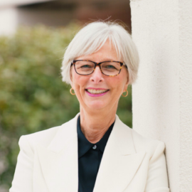 Cynthia McGuire, President & CEO of Monadnock Community Hospital, smiling while leaning against a white pillar outdoors. She is dressed in a white blazer and glasses, with greenery in the background