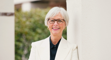Cynthia McGuire, President & CEO of Monadnock Community Hospital, smiling while leaning against a white pillar outdoors. She is dressed in a white blazer and glasses, with greenery in the background